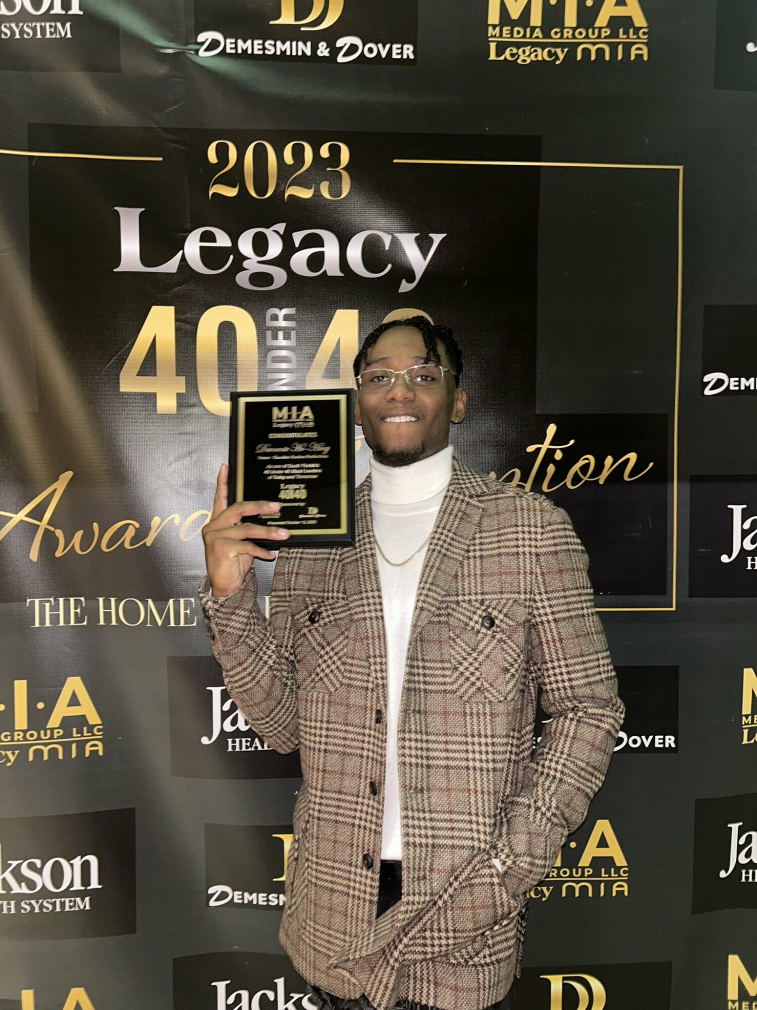 Person in plaid suit holding award plaque, standing in front of black and gold backdrop with event details.