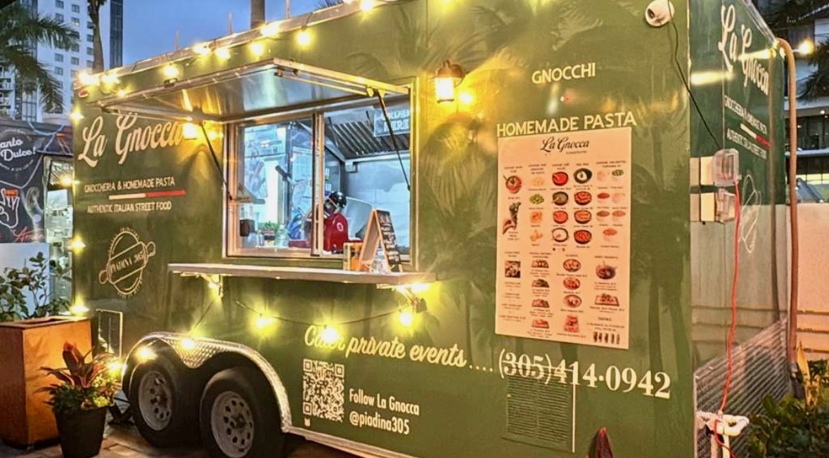 Green food truck with illuminated lights, serving pasta and gnocchi, with menu and contact information displayed on side.