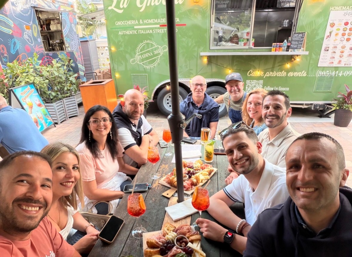 Group of people sitting around a table outdoors, enjoying food and drinks, smiling at the camera.