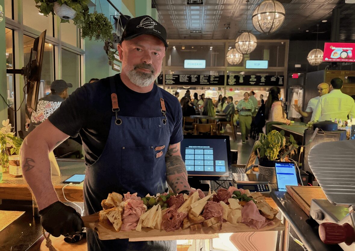 Man wearing black apron and cap stands behind a tray of sliced meat in a busy restaurant or market setting.