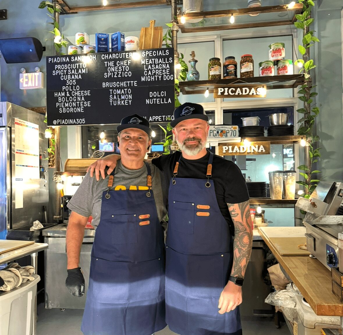 Two men wearing aprons and hats stand inside a food stall, smiling with arms around each other.