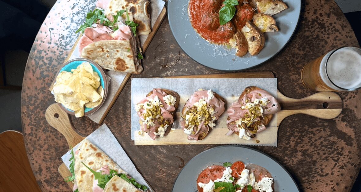 Assorted dishes including sandwiches, appetizers, and drinks on a round wooden table.