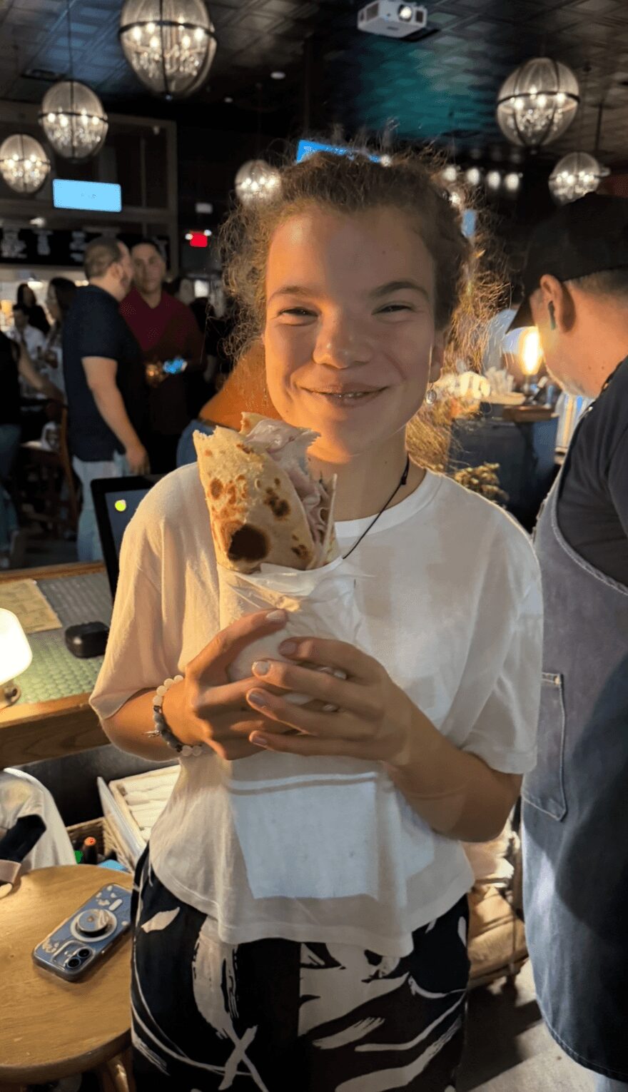 Smiling girl holding a slice of cake in a lively indoor setting with hanging lights.