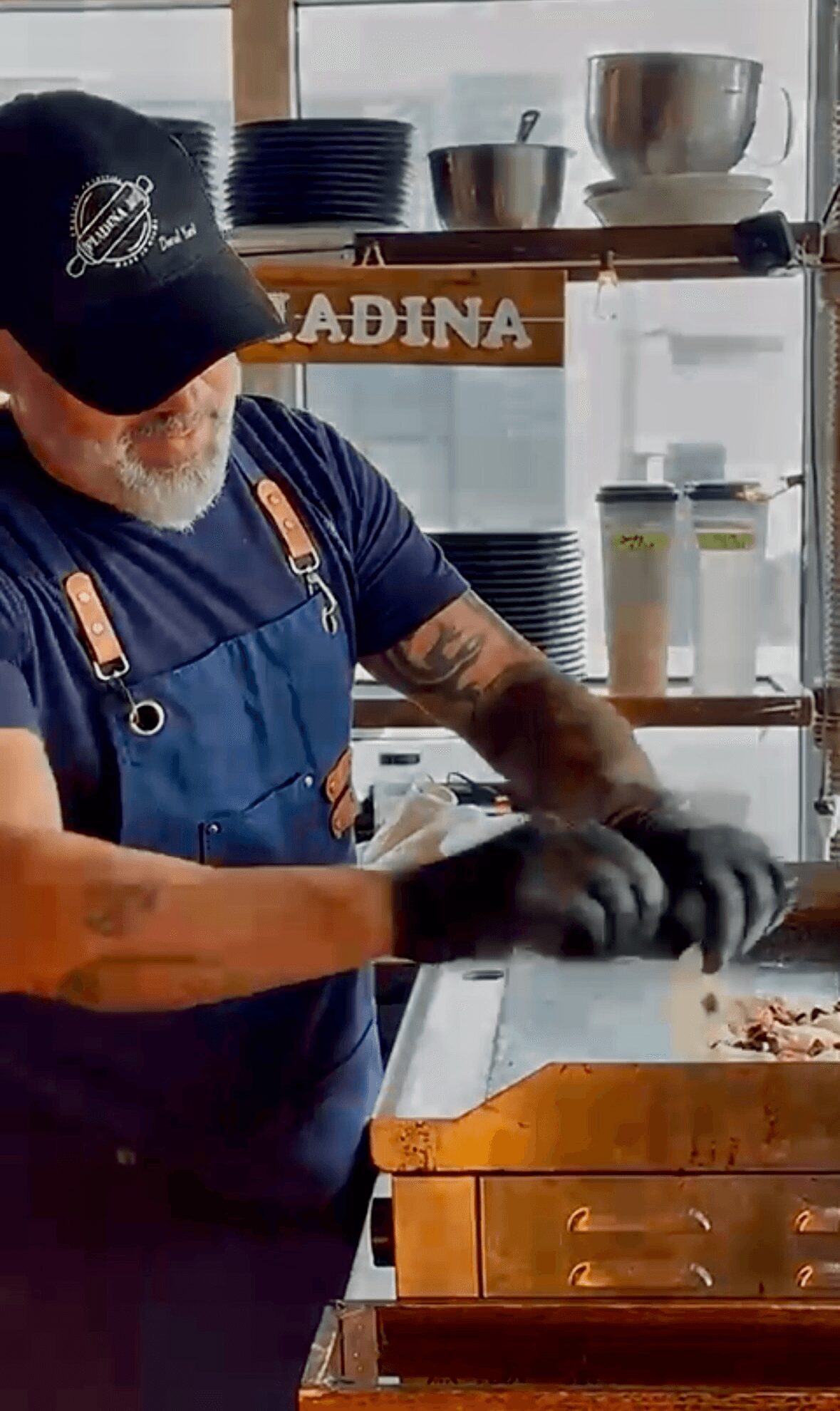 Person wearing a black cap and apron, working with food in a kitchen setting.