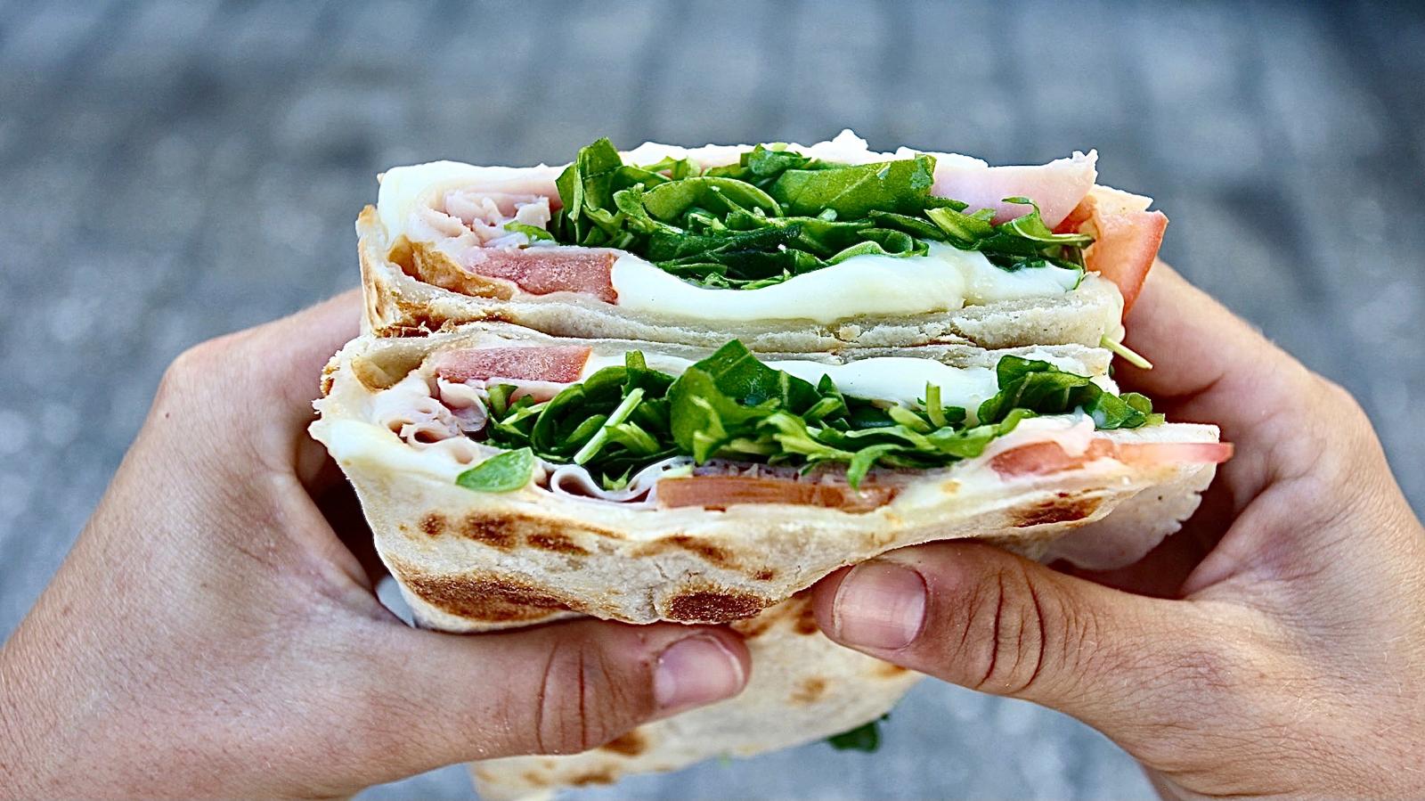 Person holding a sandwich with layers of meat, cheese, and green vegetables, in a hand against a blurred background.