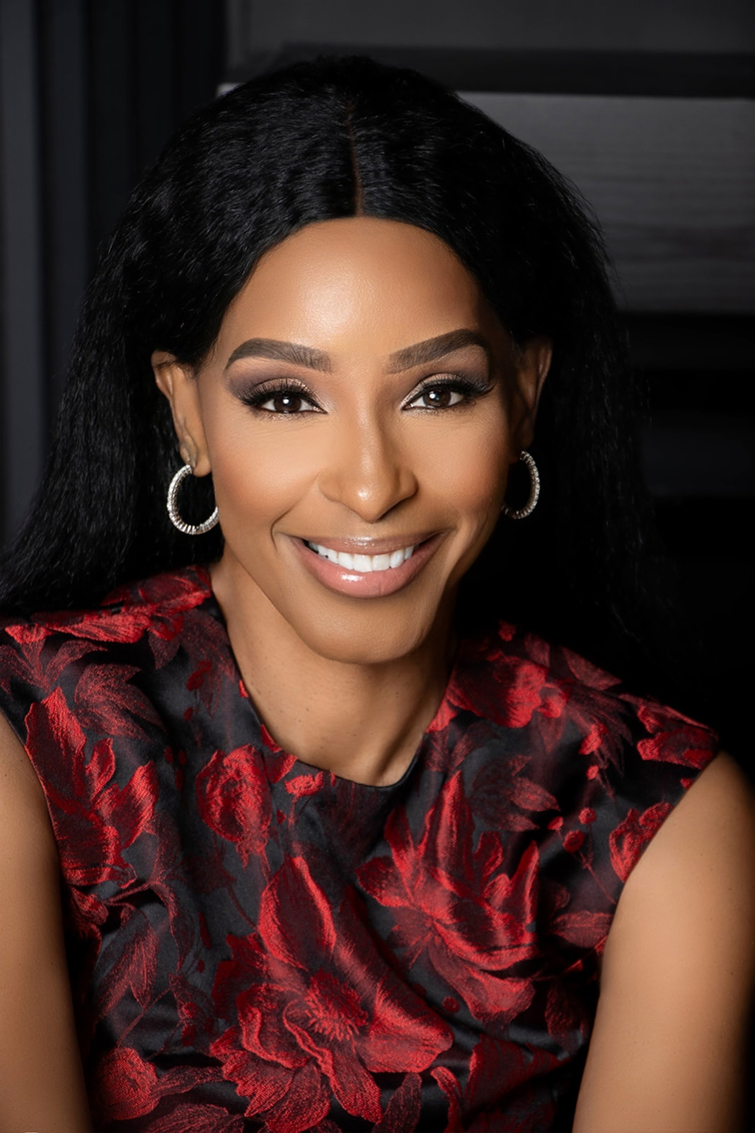 Smiling woman with dark hair, hoop earrings, wearing a red and black patterned top.