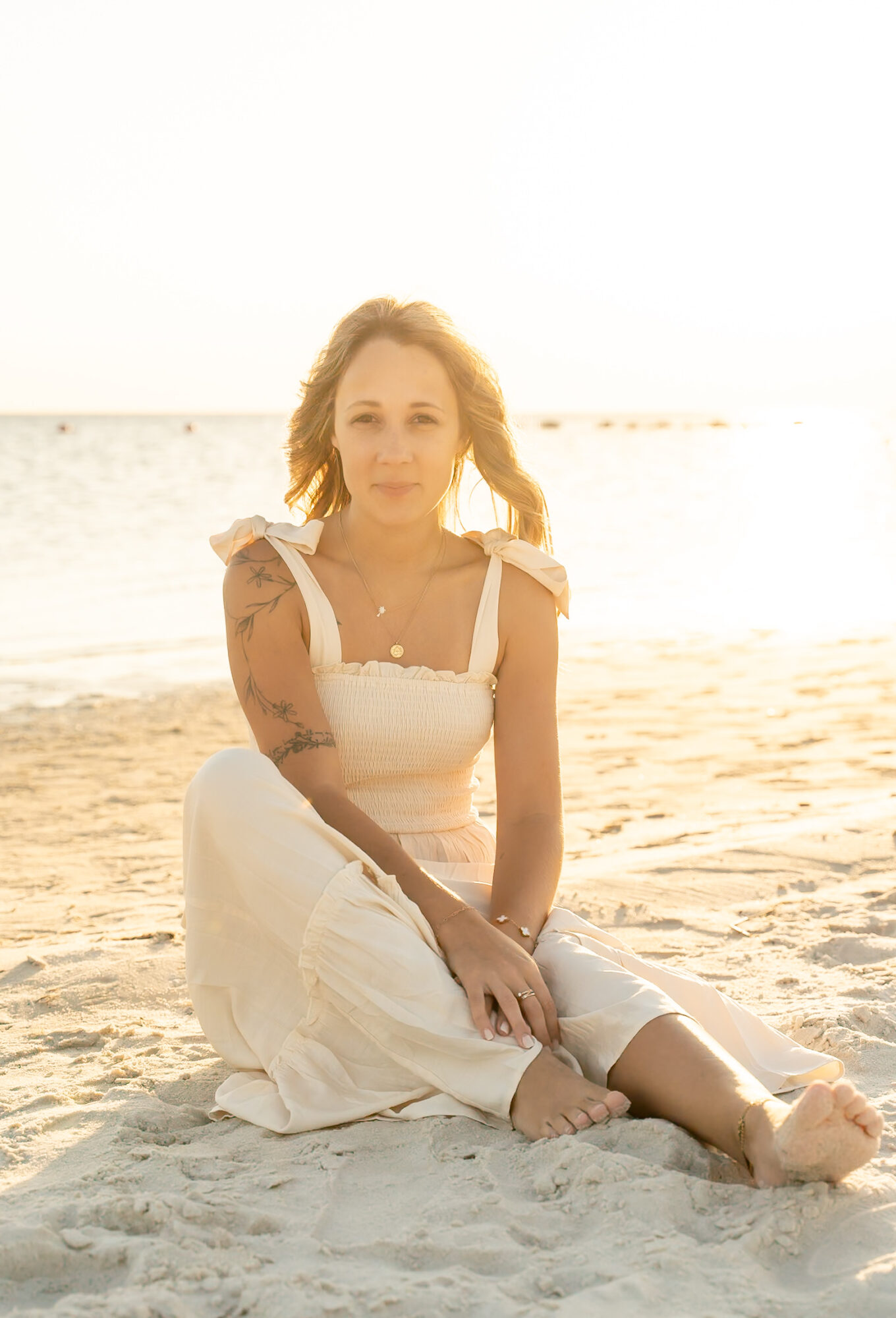 Woman sitting on sandy beach with ocean in background, wearing a white dress and smiling.