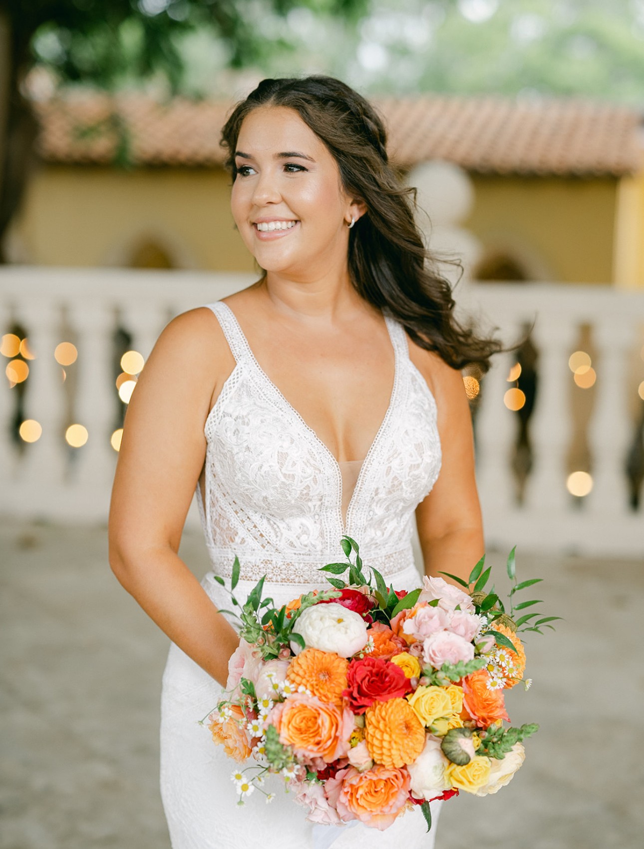 Woman in white dress holding a colorful bouquet outdoors, smiling, with blurred background of trees and building.
