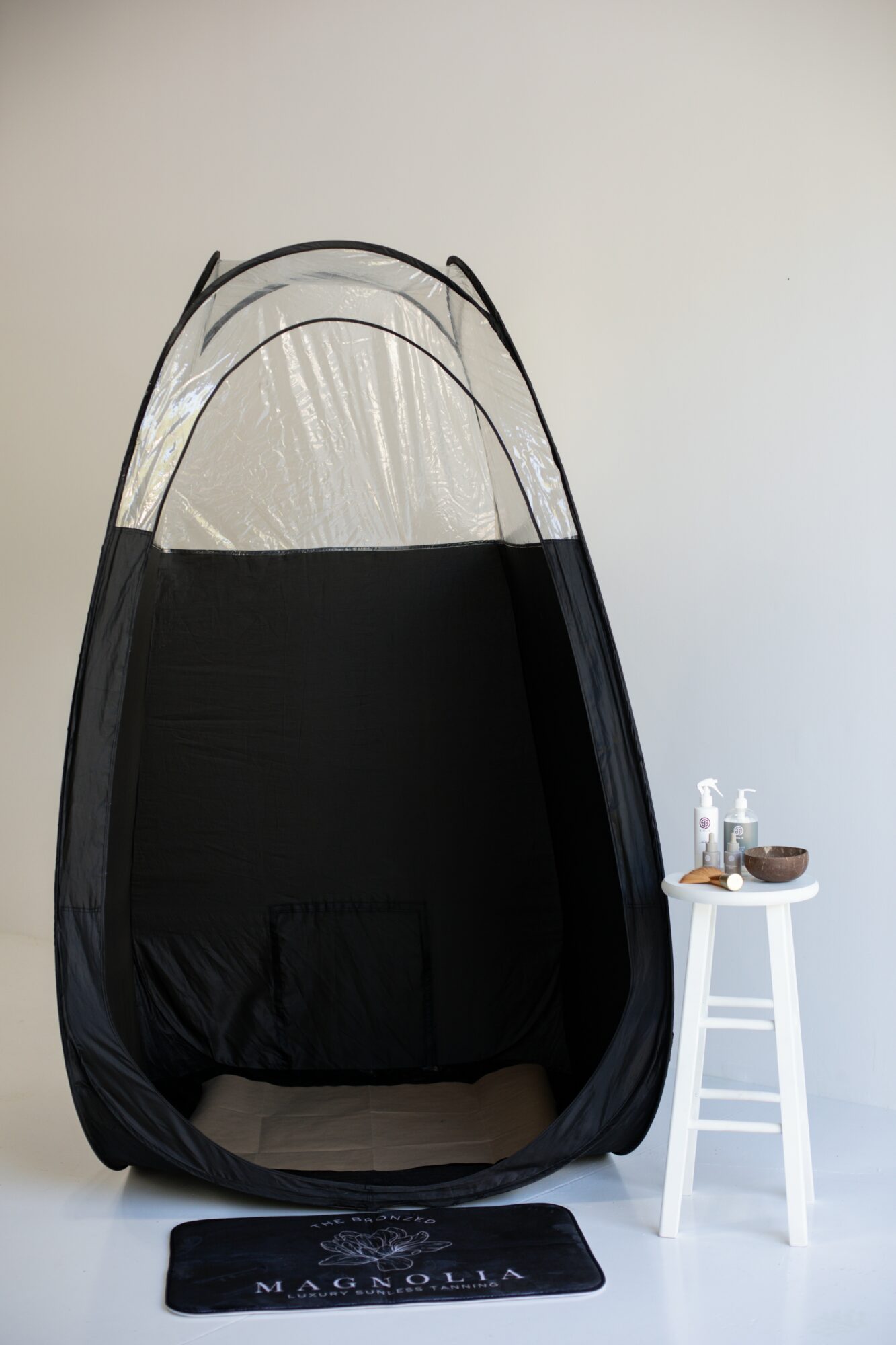 Indoor black hanging tent with a transparent top, next to a white stool with a small bottle and bowl, on a white floor.