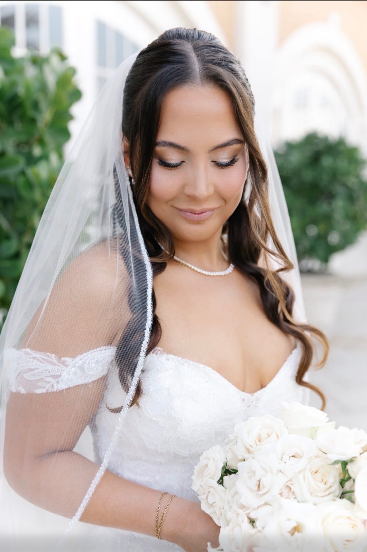 Bride with long dark hair, wearing a white wedding dress and veil, holding a bouquet of white roses outdoors.