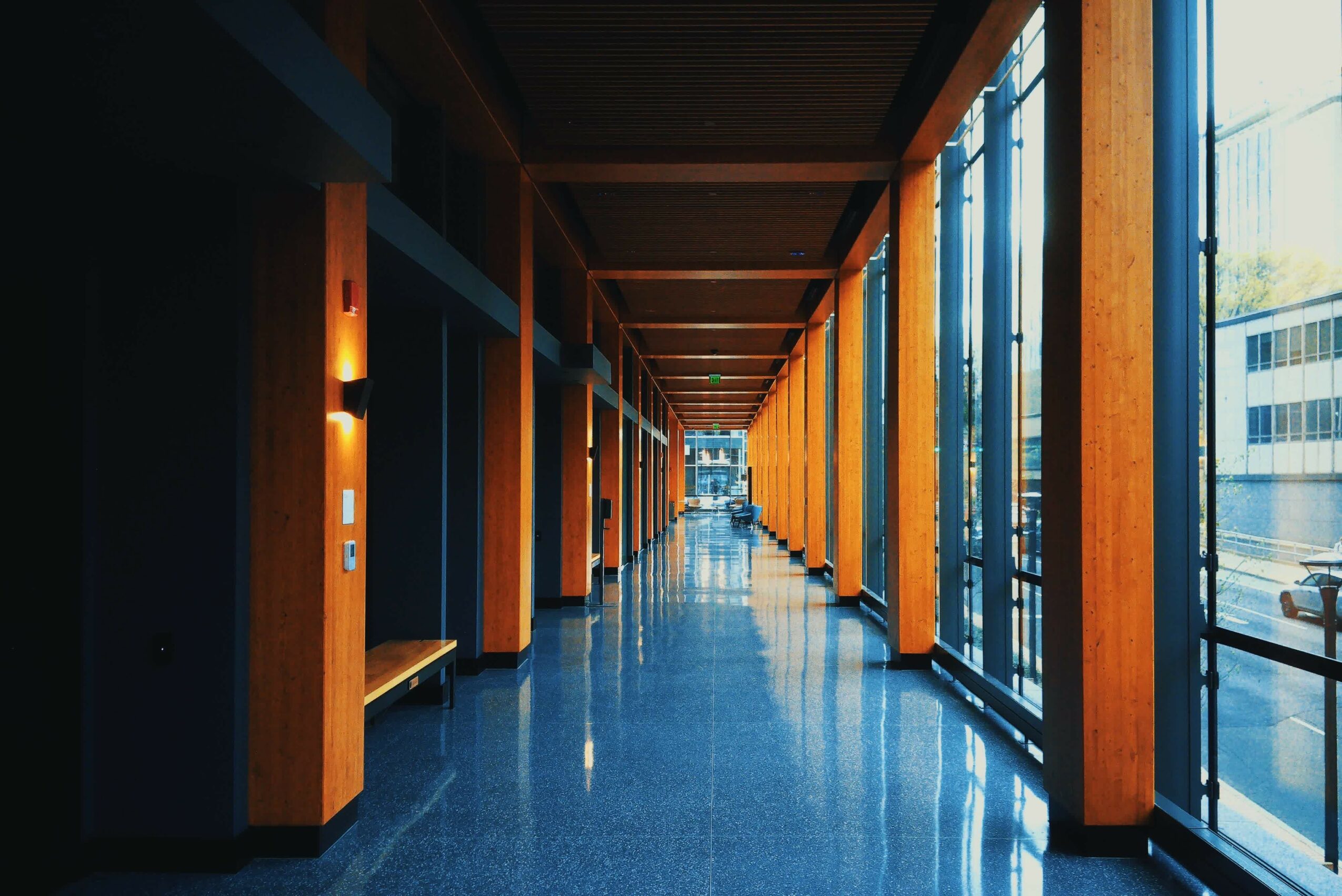 Long corridor with wooden beams, large glass windows, and a shiny blue floor, leading to a distant door.