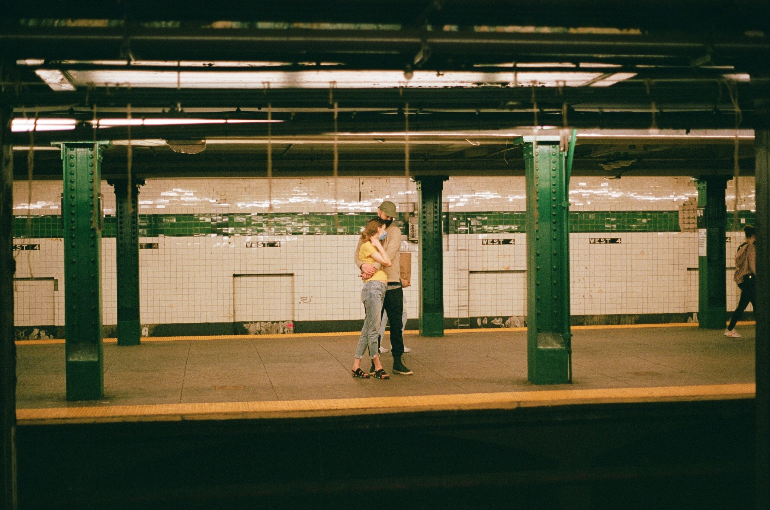 Two people embrace on a subway platform with green pillars and tiled walls in the background.