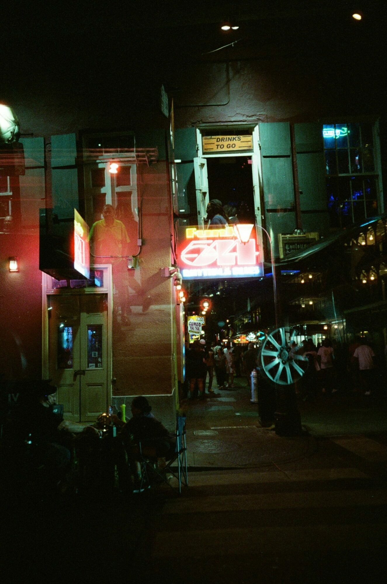 Night scene of a street with illuminated signs, buildings, and parked cars, including a large wheel and a neon sign.