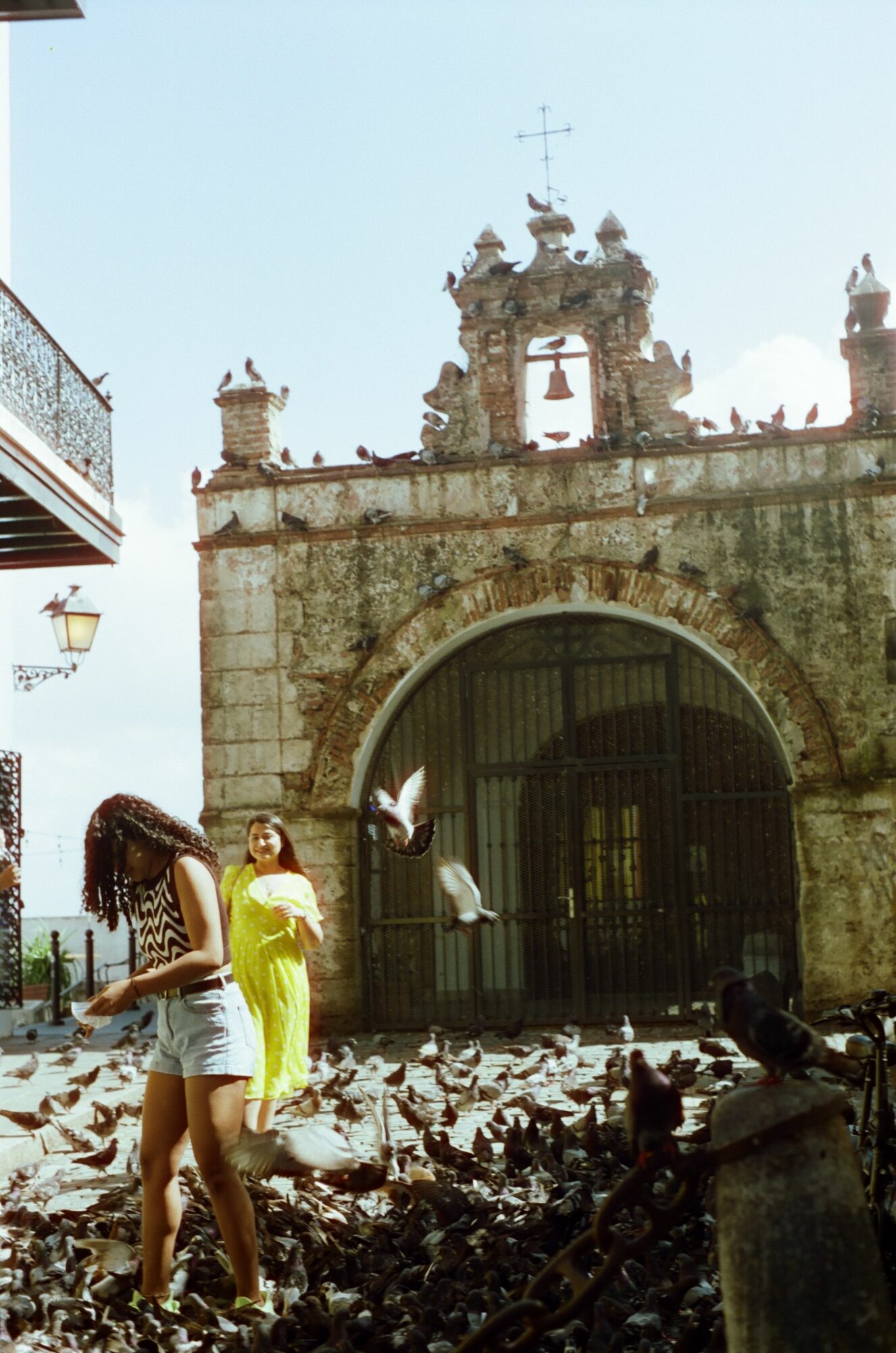 Two women near a stone archway with pigeons on the ground and a historic building in the background.