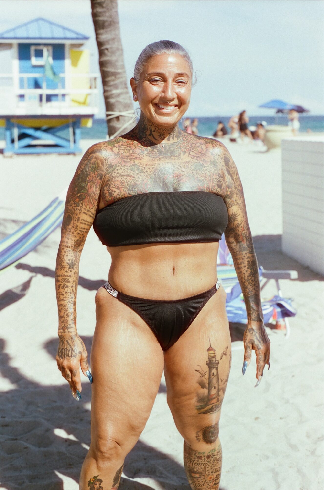 Smiling woman in black bikini standing on beach with lifeguard tower and umbrellas in background.