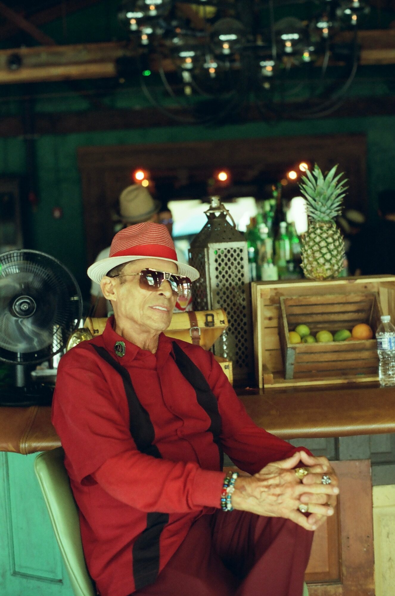 Older woman wearing a red hat and sunglasses, sitting with hands clasped, in a colorful indoor setting with plants and decorations.