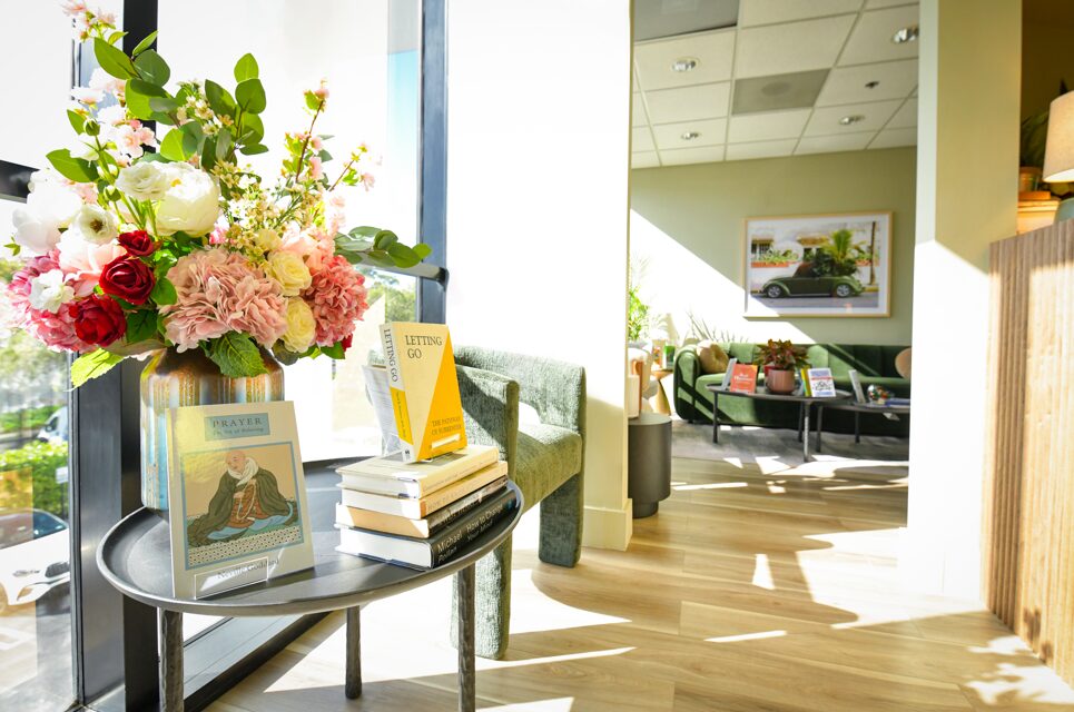 Table with a large flower arrangement and books inside a bright room with seating area and large windows.