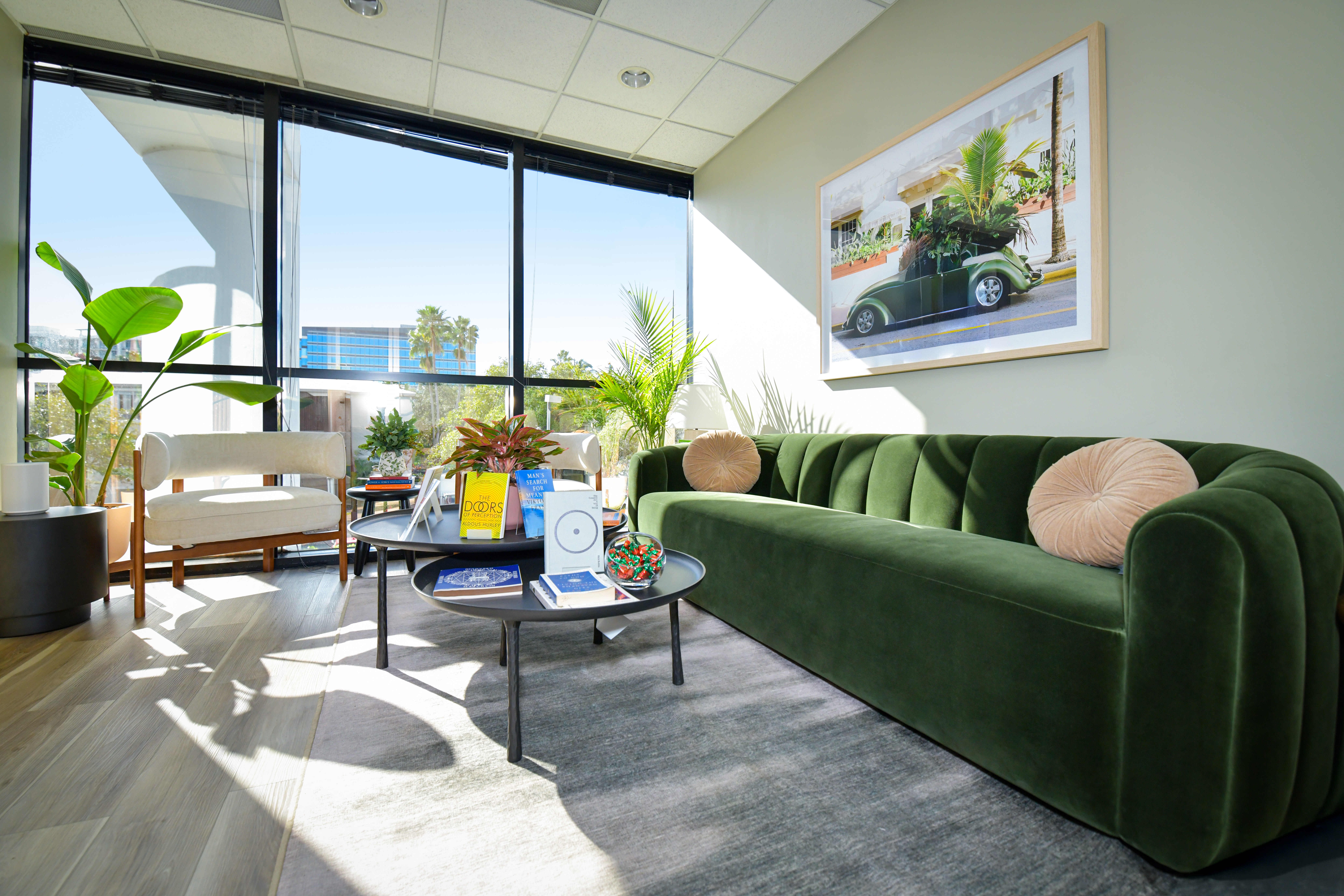 Modern lounge with green velvet sofa, round coffee tables, large window, and artwork on wall.