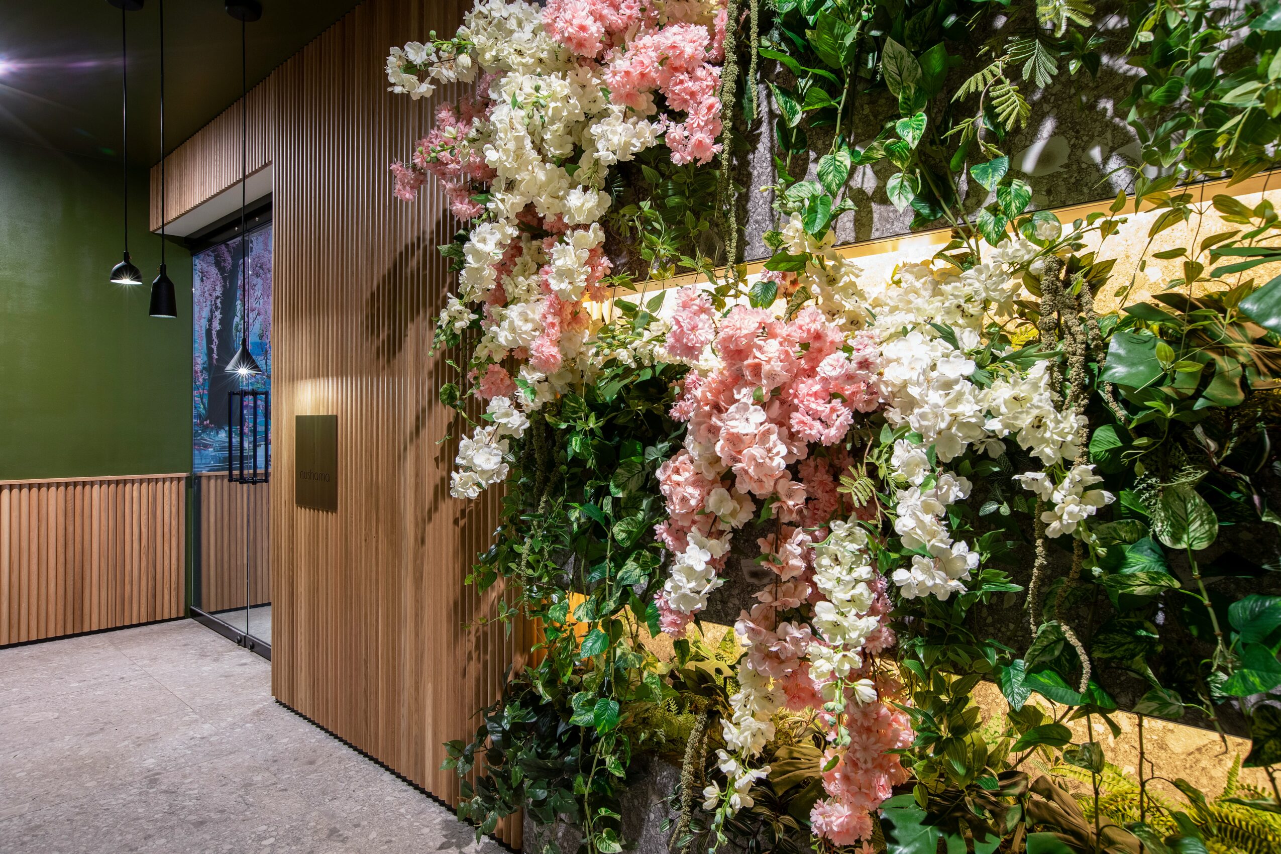 Close-up of pink and white flowers with green leaves on a wall, indoors with wooden paneling and a window in the background.