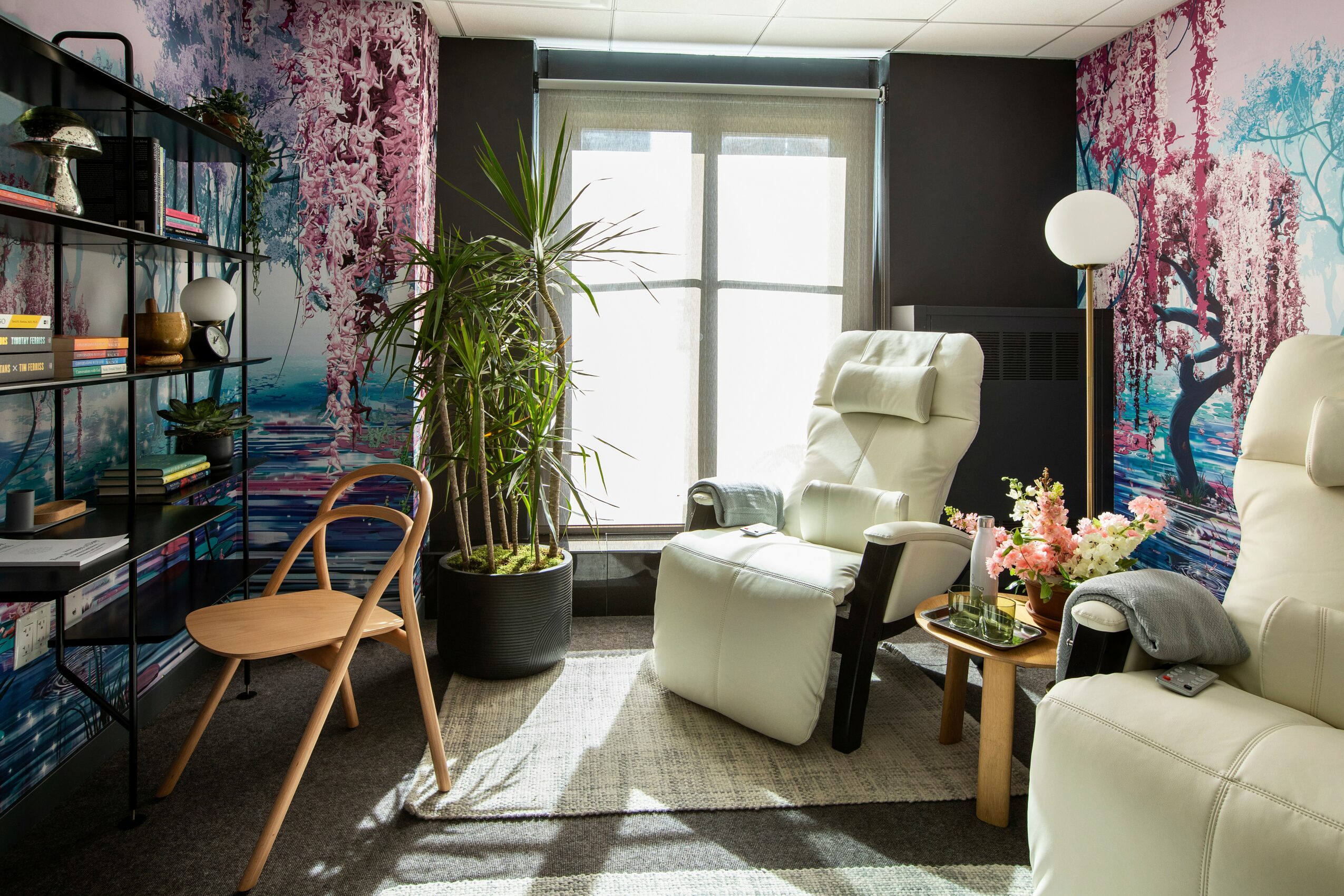 Room with two recliner chairs, a tall plant, a side table with flowers, and a bookshelf, window in the background.