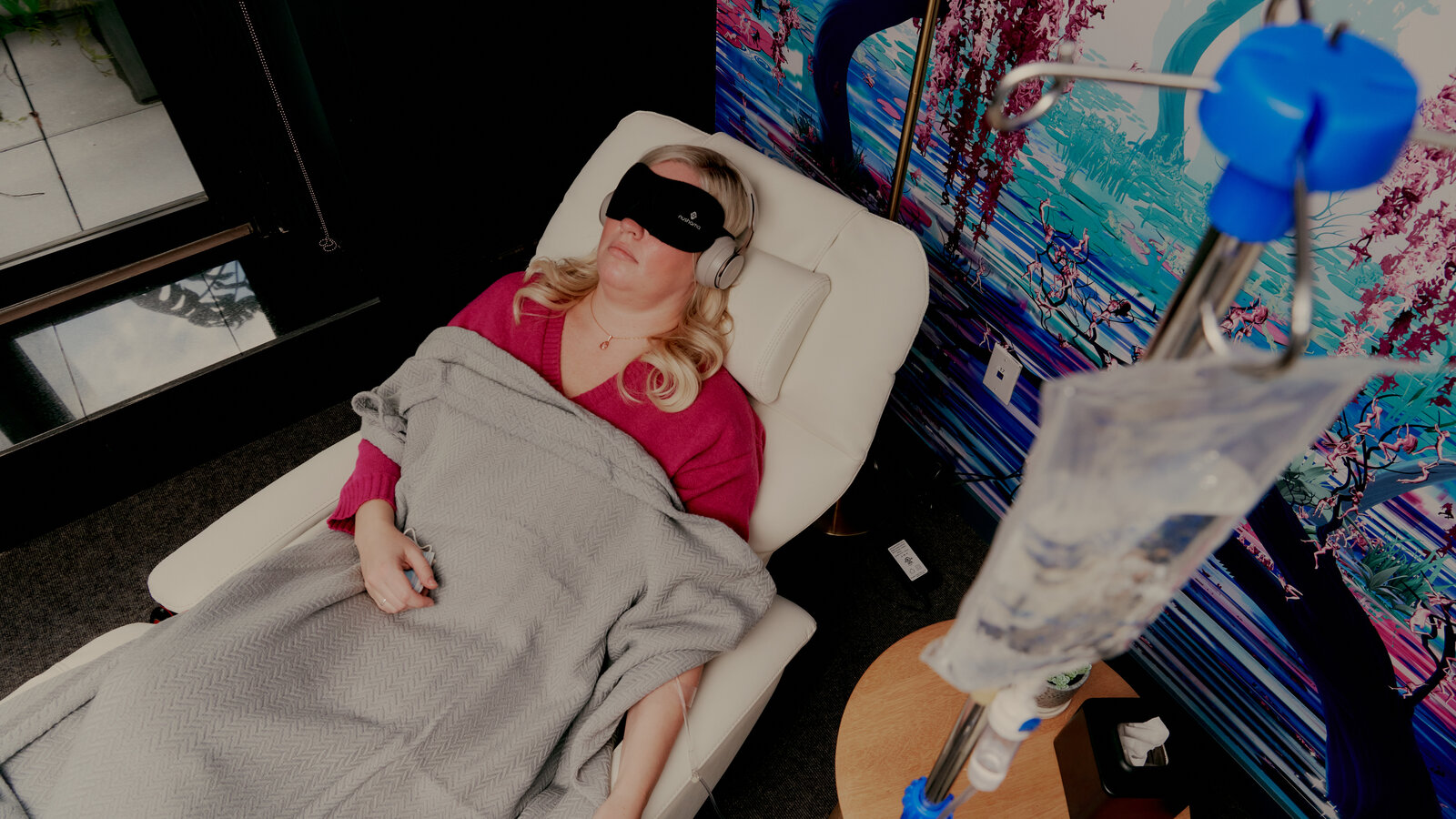 Person lying in a hospital bed with an IV drip, wearing a blindfold and a red top, covered with a blanket.