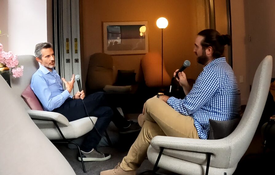 Two men sit in chairs facing each other, engaged in conversation in a cozy room with warm lighting.