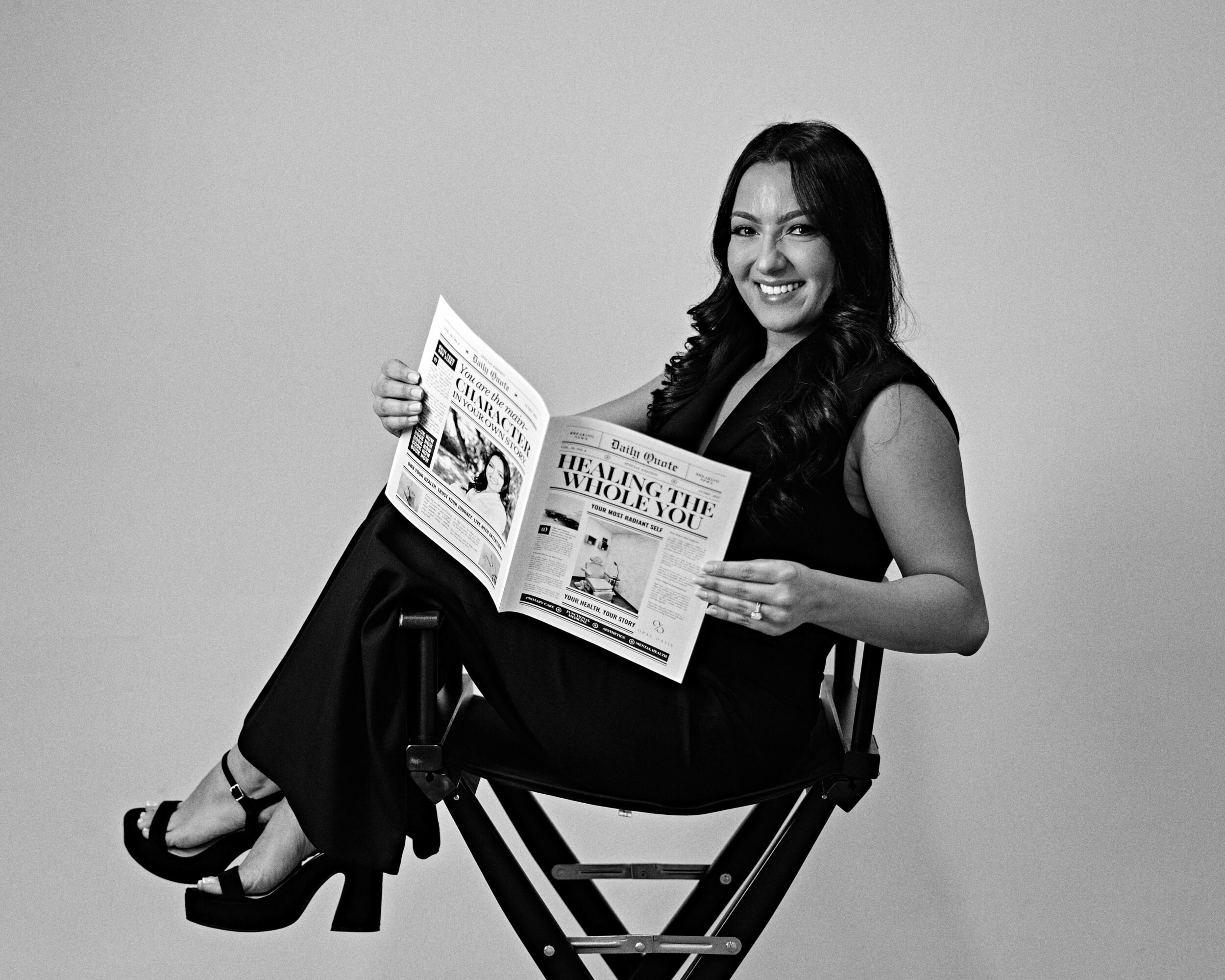 Woman sitting on a director's chair, smiling, reading a newspaper, in black and white.