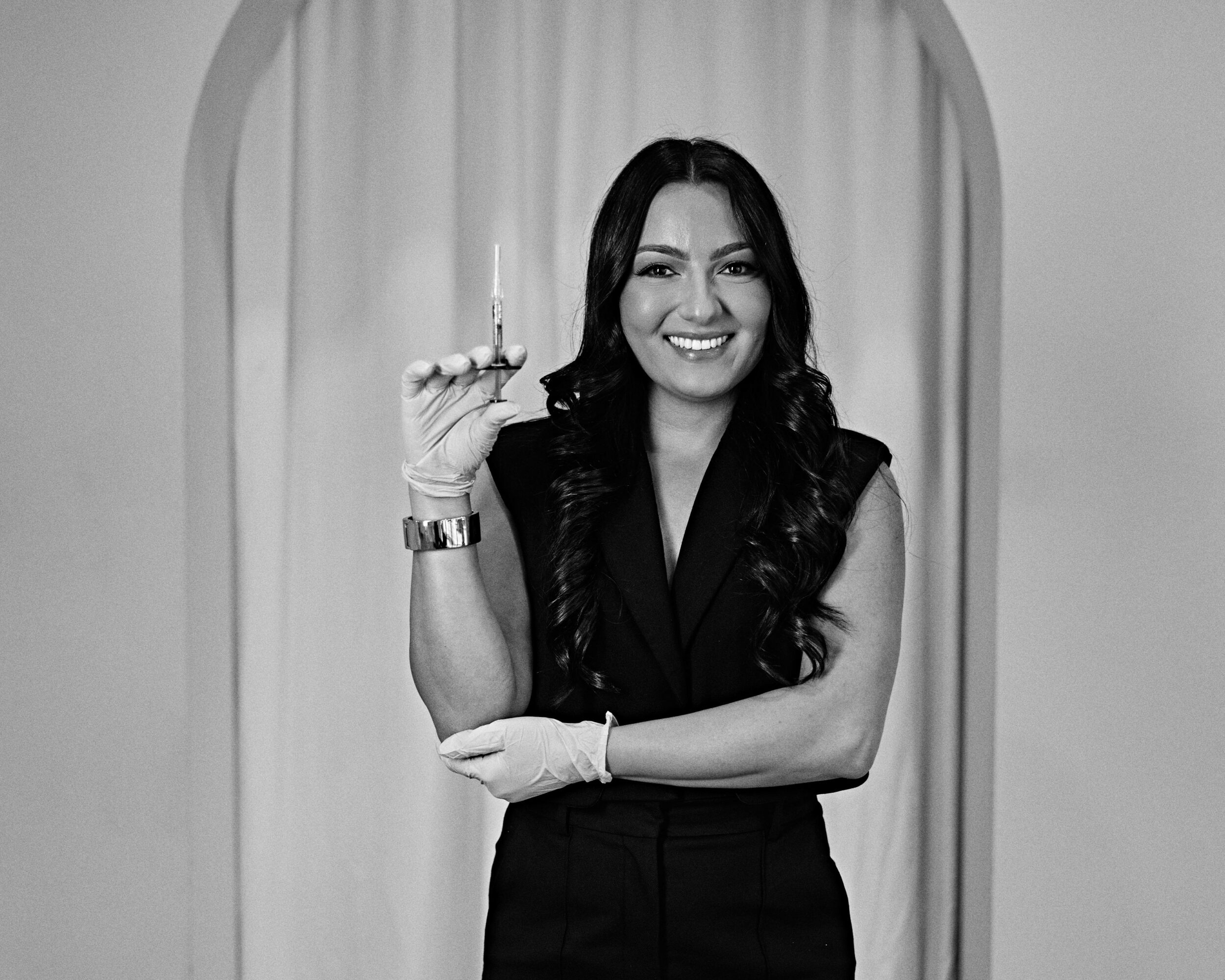 Woman with long dark hair smiling, holding a lit candle, standing in front of curtains and an archway.