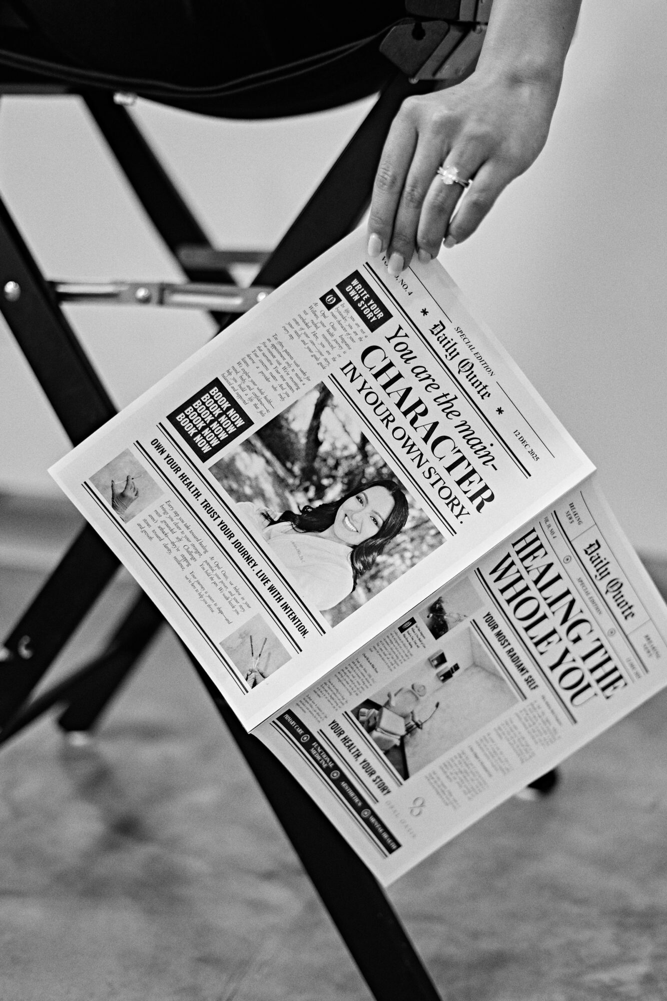Person's hand holding a newspaper, with a chair and ground in the background.