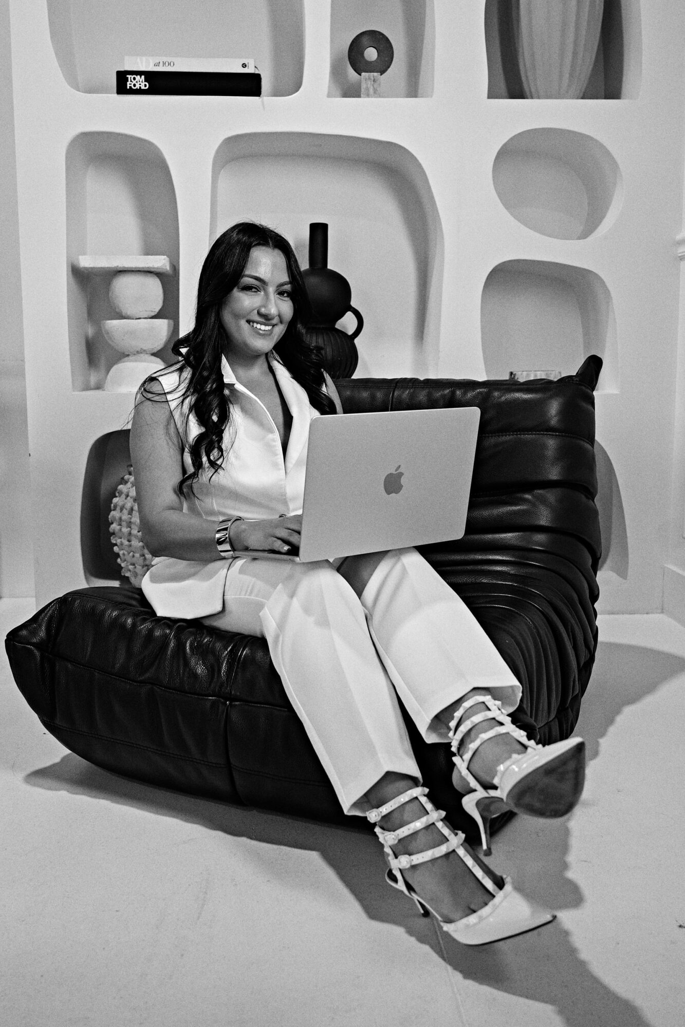 Woman sitting on a black bean bag chair with a laptop, smiling, in a modern room with shelves and decorative objects.