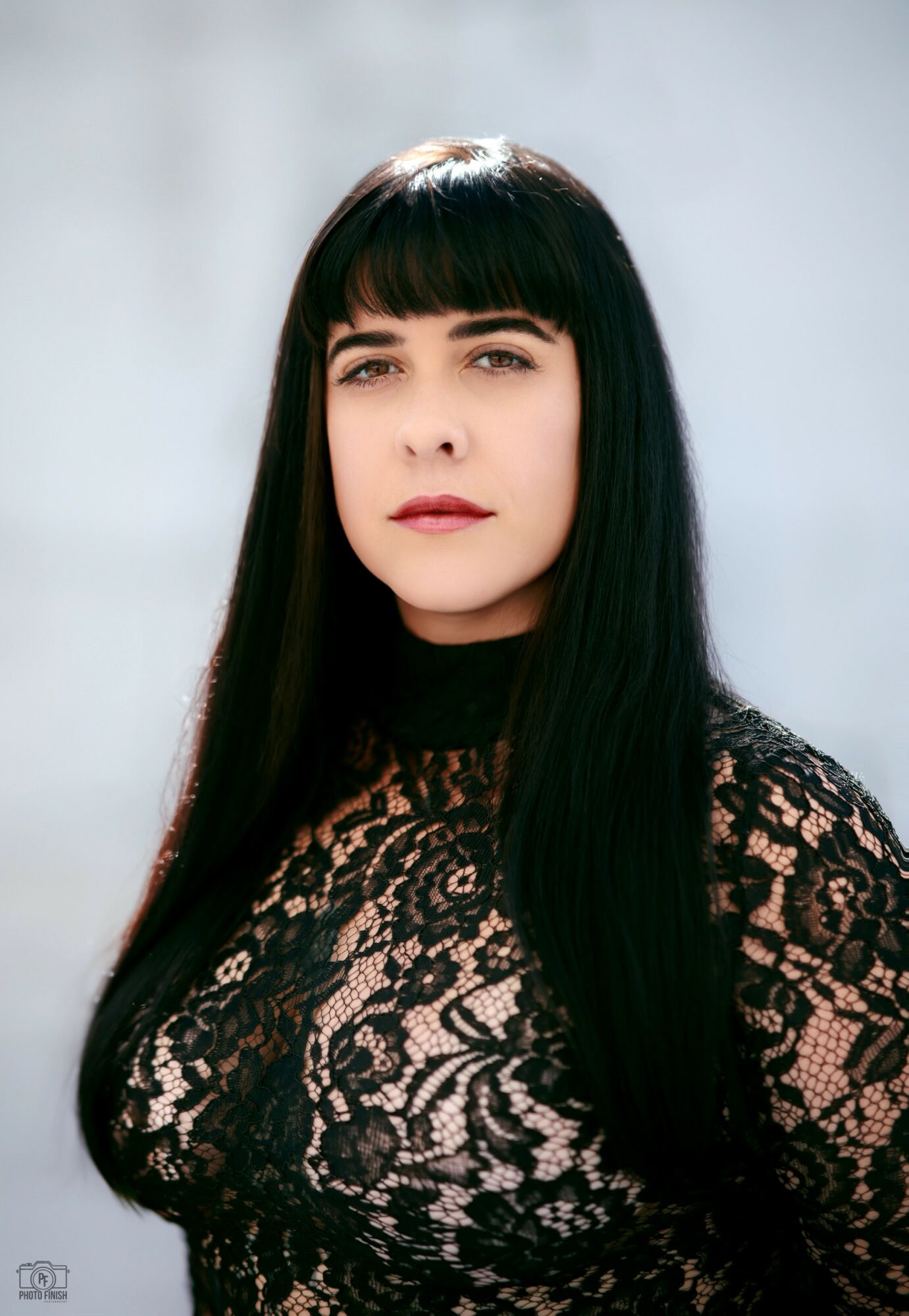 Woman with long black hair and bangs wearing a black lace top, looking at the camera outdoors.