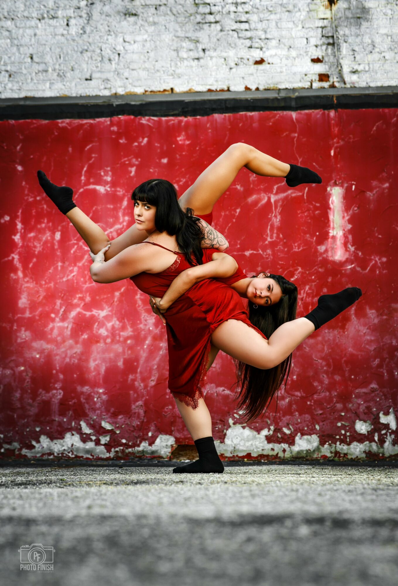 Two women in red dresses and black shoes perform a dance pose against a red textured wall.