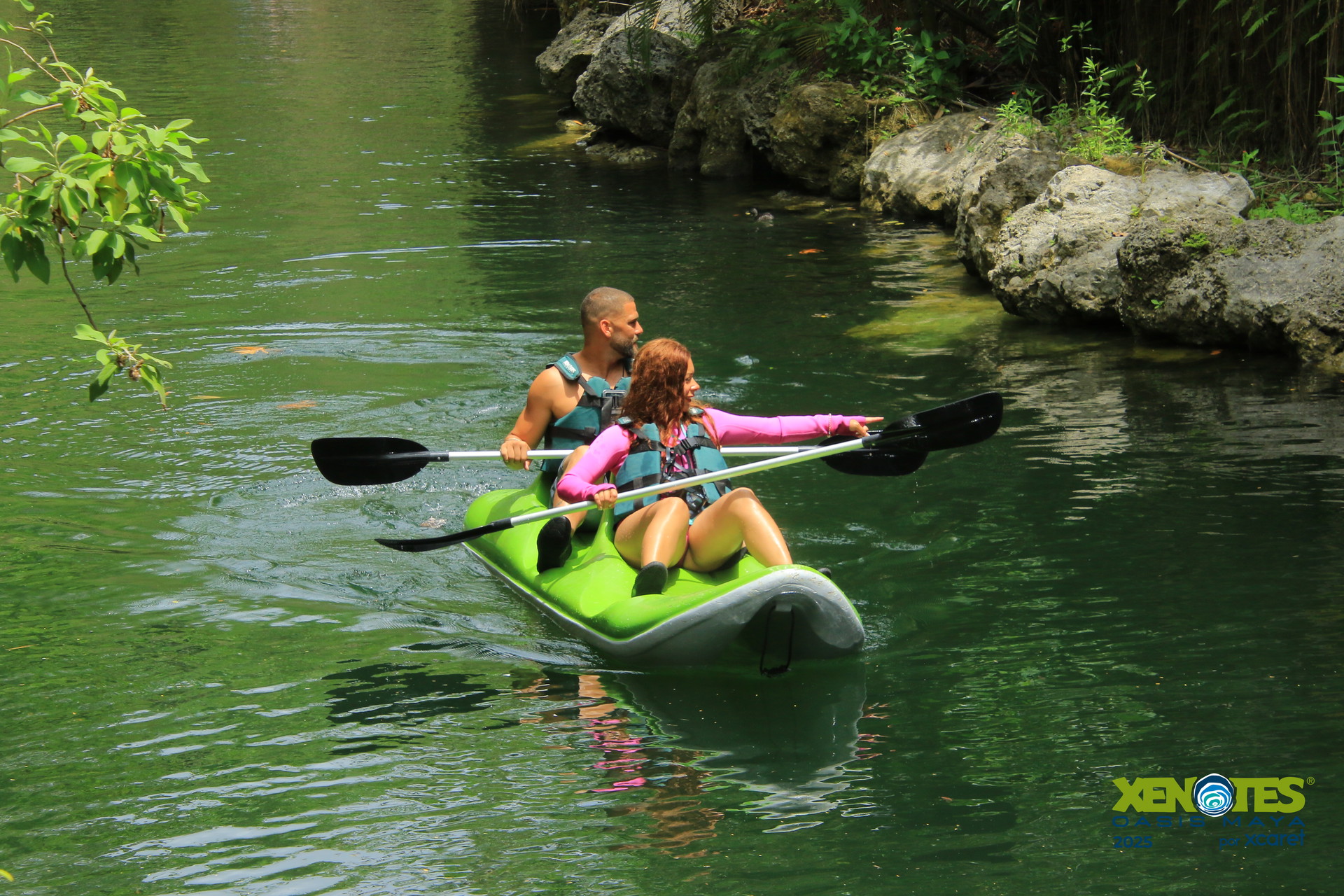 Two people in a green kayak on a calm river, surrounded by rocks and greenery, with one person paddling and the other sitting.