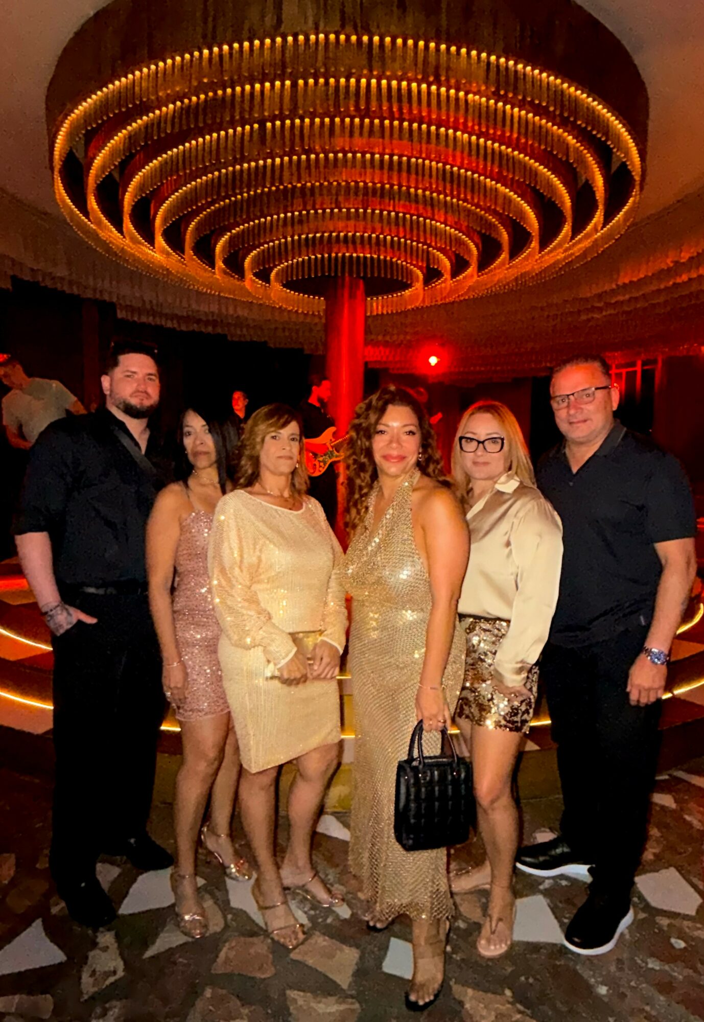 Group of six people standing in a decorated venue with a large circular chandelier overhead.
