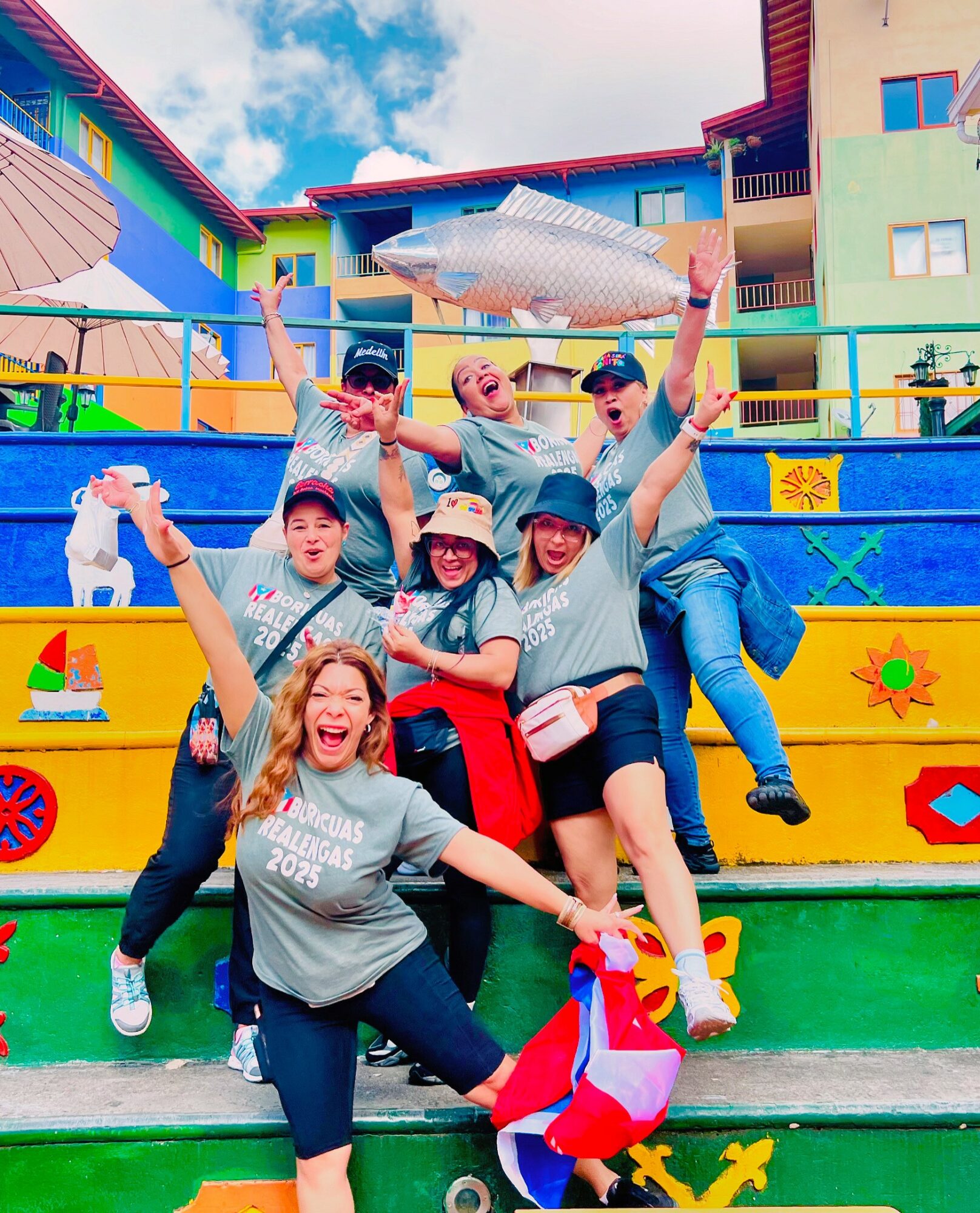 Group of six people on colorful outdoor stairs, smiling, holding a large fish-shaped object, with buildings and sky in background.