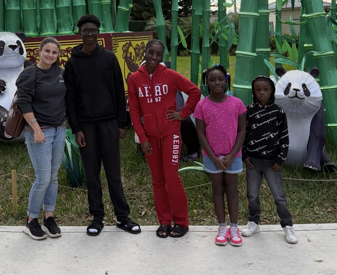 Group of five children standing outdoors in front of panda statues and bamboo decorations.