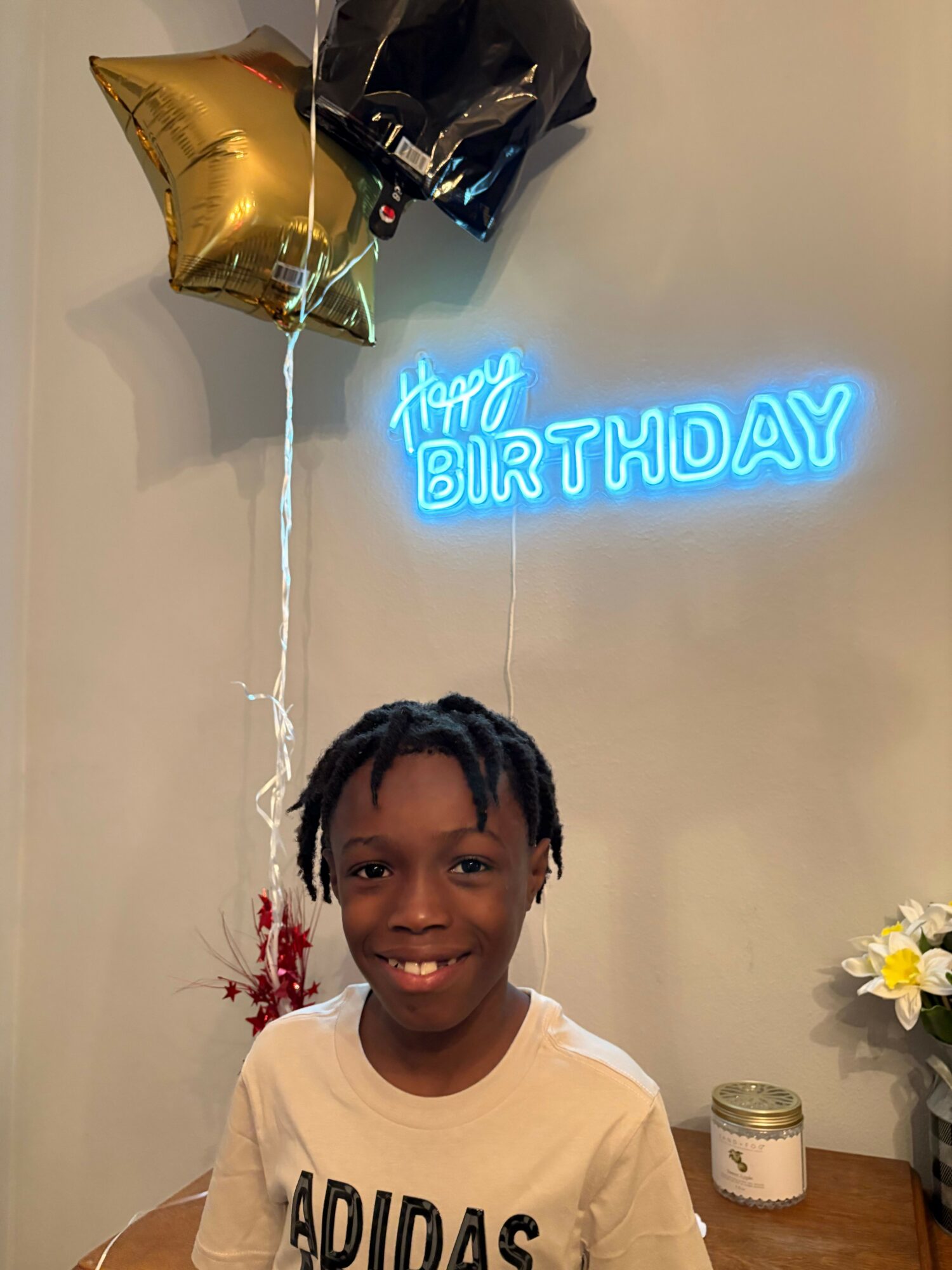 Child with short hair smiling in front of balloons and a birthday sign on wall.