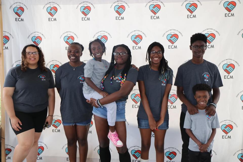 Six people standing in front of a backdrop with logos, smiling, with one child being held.