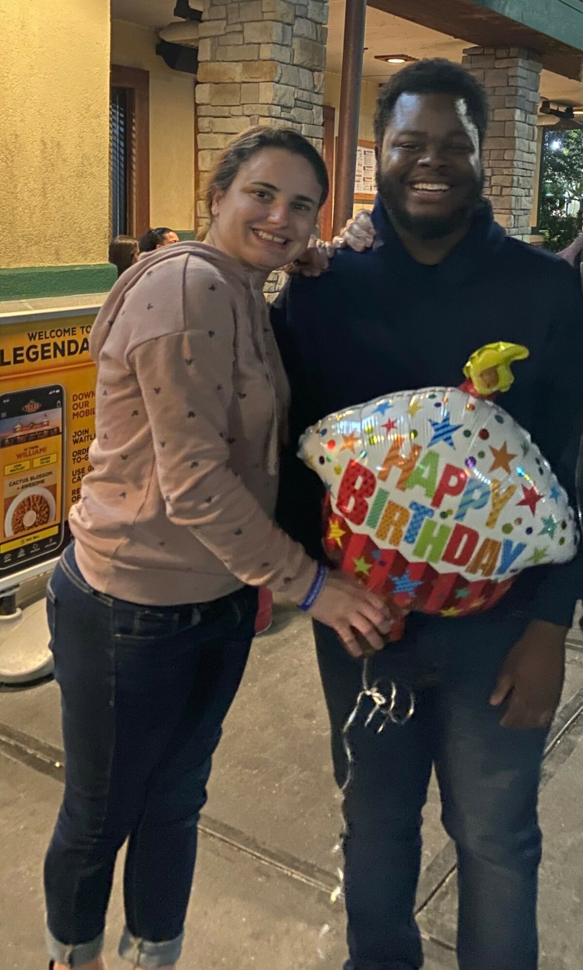 Two people smiling, holding a birthday balloon, standing outside near a yellow and brick building.