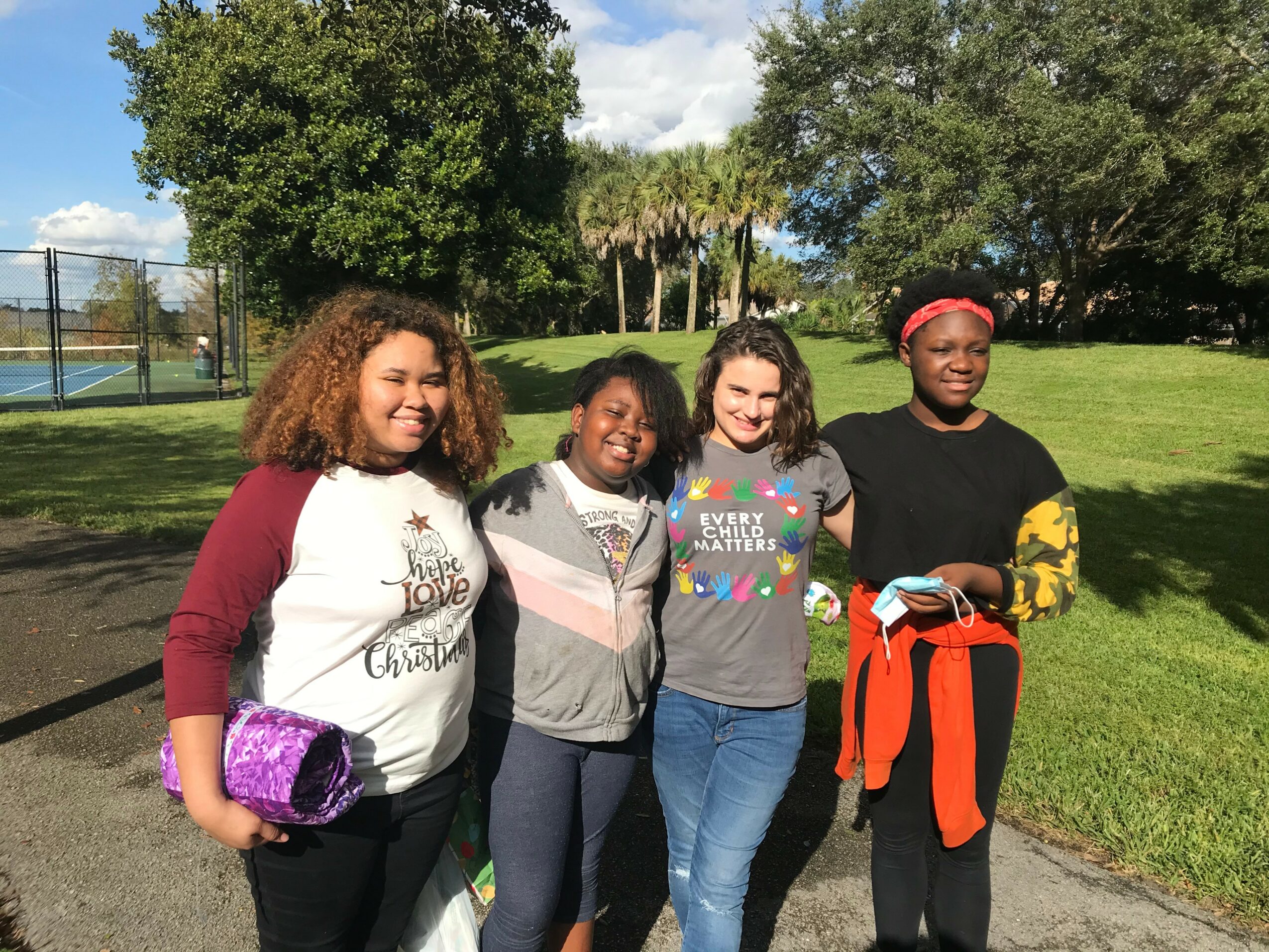 Four women standing outdoors on grass, smiling, with trees and a fence in the background.