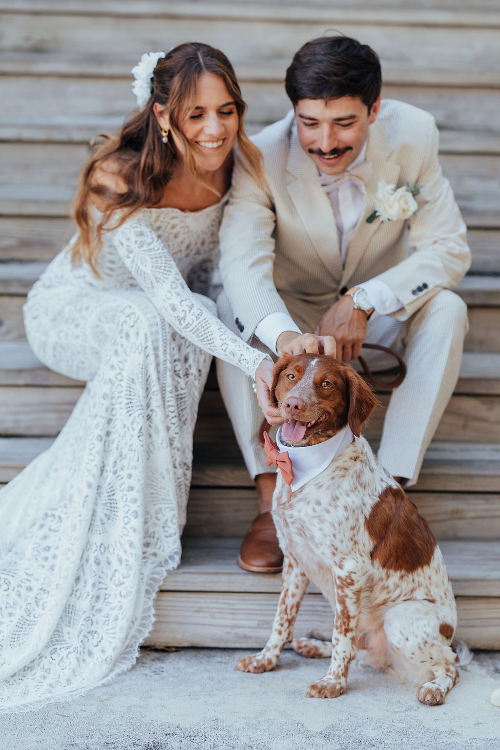 Bride and groom smiling with a dog on steps, bride in white dress, groom in light suit, dog with pink bandana.