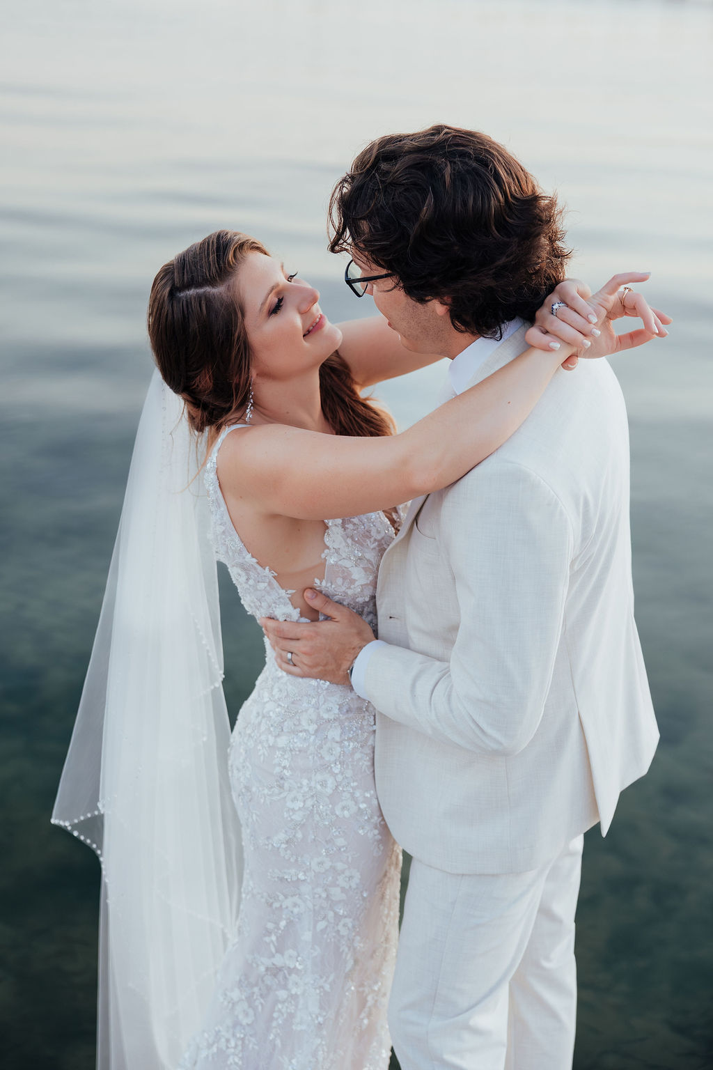 Bride and groom embrace near water, gazing into each other's eyes, with the bride in a lace wedding dress and veil.