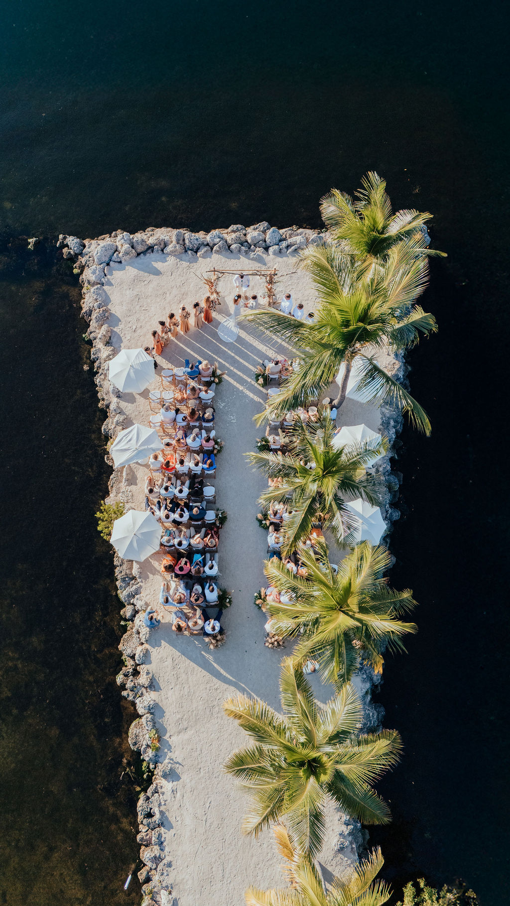 Beachside setup with umbrellas, lounge chairs, and palm trees on a narrow strip of land surrounded by water.