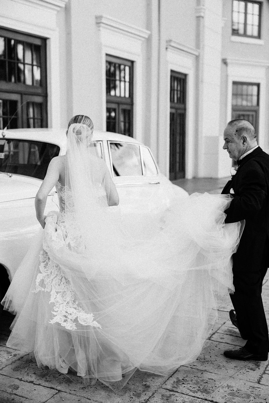 Bride in wedding dress and veil, man in suit, outside building with large windows and a vintage car.