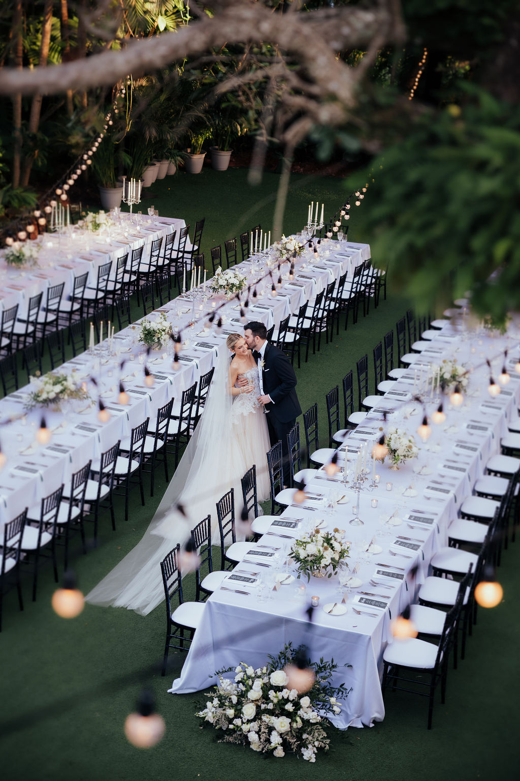 Wedding reception setup with long tables, floral centerpieces, and a bride and groom standing together.