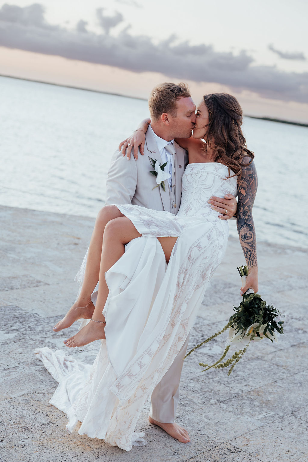 Couple on beach, man carrying woman in wedding dress, holding bouquet, sunset sky in background.