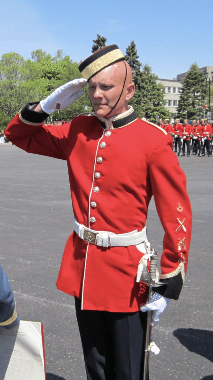 Military officer in red uniform saluting outdoors with a group of soldiers in the background.