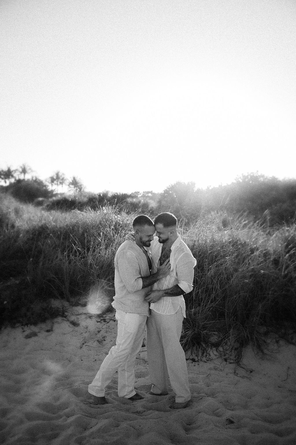 Two people stand close together on a sandy beach, holding hands and looking at each other, with grassy dunes in the background.