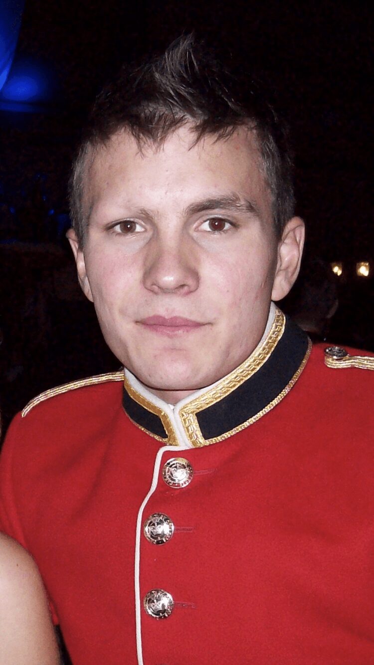 Young man in a red military-style uniform with gold accents and buttons, dark background, looking at camera.