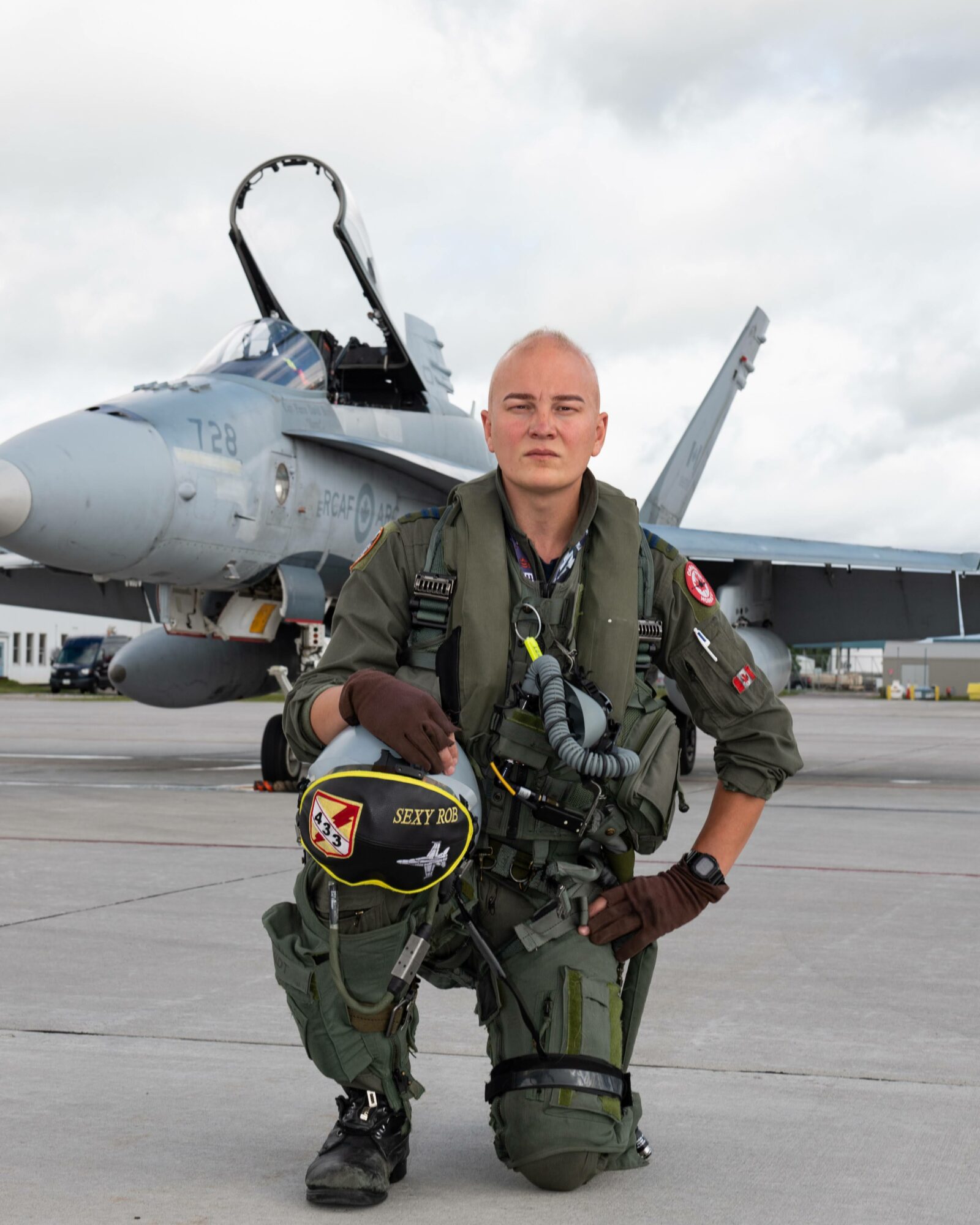 Female pilot in flight suit kneeling on tarmac with fighter jet in background under cloudy sky.